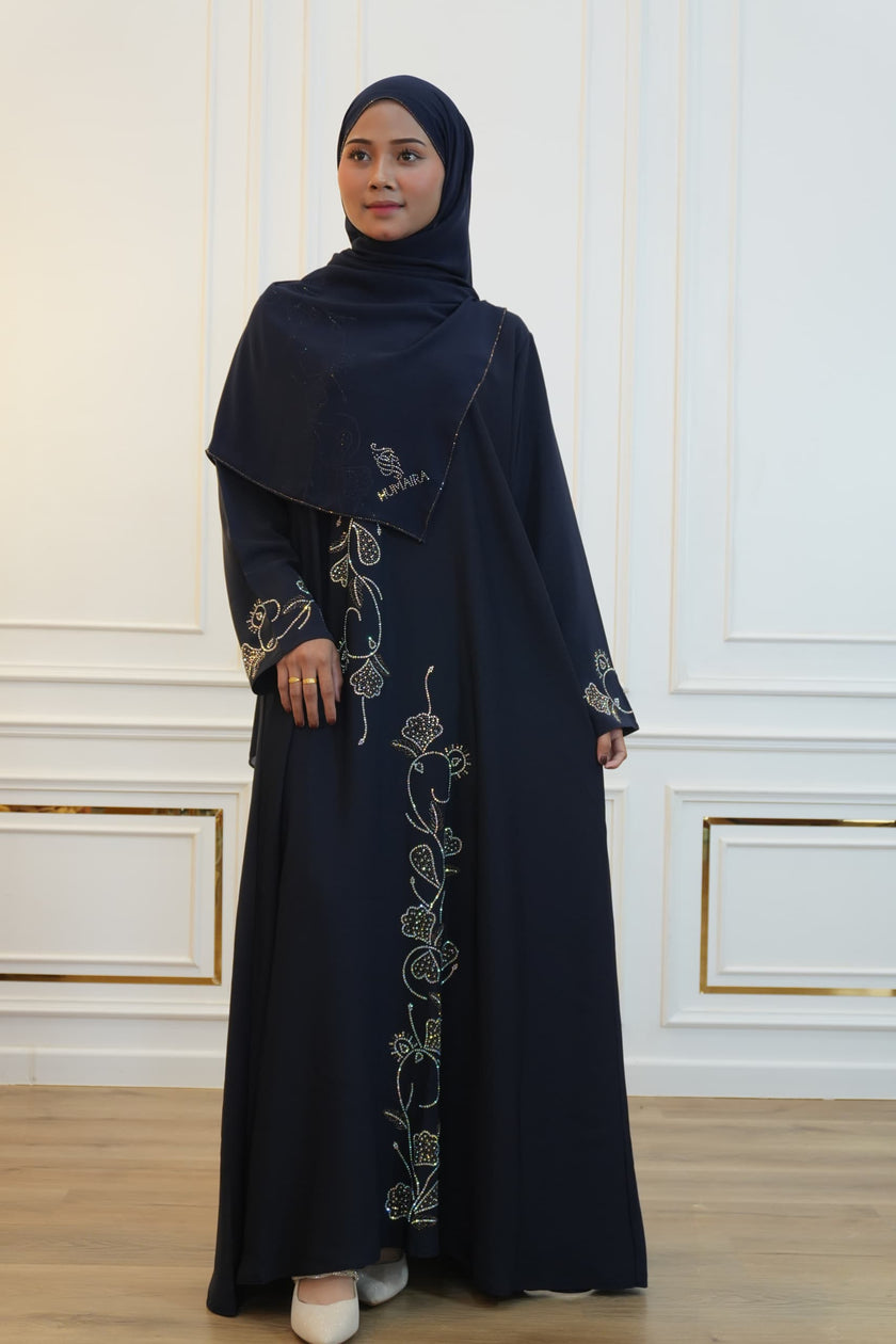 Woman wearing a black abaya with floral embroidery in a room with white walls and wooden floor.